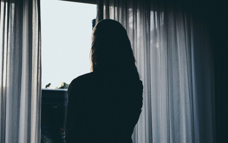 Silhouette einer Person, die durch halbgeöffnete Vorhänge nach draußen blickt und die Dämmerung beobachtet.