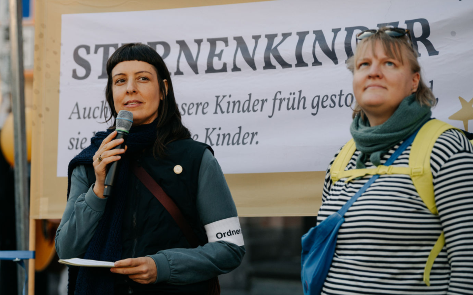 Jessica Hefner spricht in ein Mikrofon, während eine andere Claire Lerner, gekleidet mit gestreiftem Shirt und Rucksack, zuhört. Im Hintergrund ein Banner mit der Aufschrift 'STERNENKINDER'.