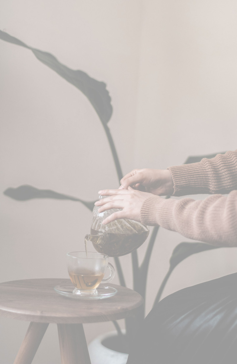 Eine Person gießt Tee aus einer Glaskanne in eine Tasse auf einem kleinen Tisch.