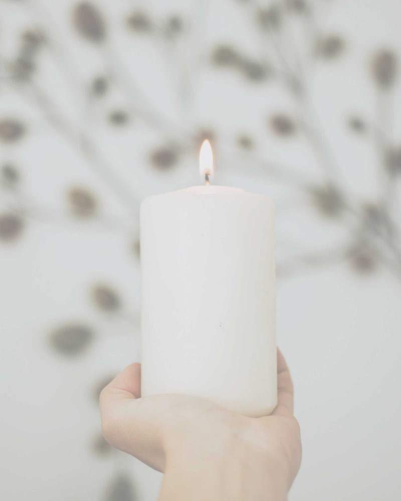 Eine Hand hält eine brennende weiße Kerze vor hellem, ruhigem Hintergrund.