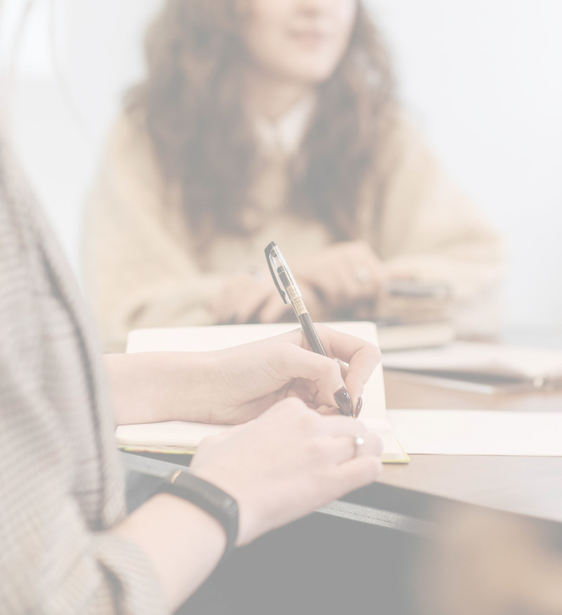 Eine Person schreibt Notizen in ein Heft, im Hintergrund sitzt eine weitere Person an einem Tisch.