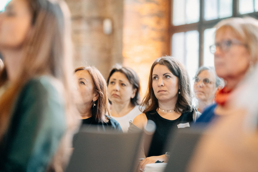 Publikum hört aufmerksam einem Vortrag zu, Fokus auf einzelne Gesichter im Vordergrund.